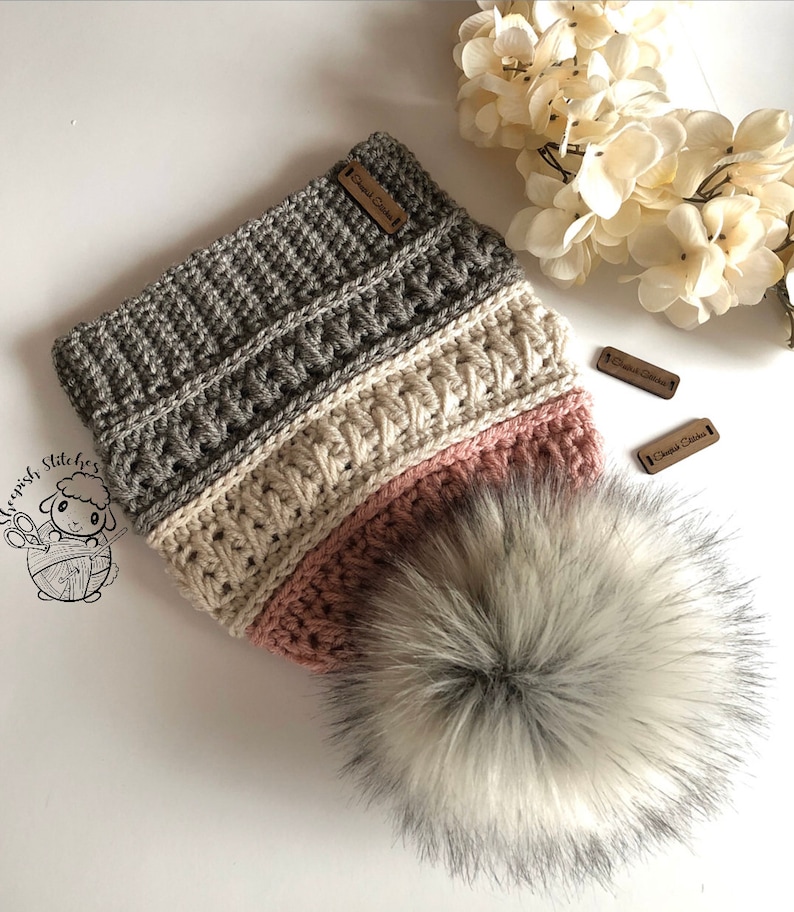 May include: A gray, cream, and pink crocheted beanie with a fluffy white pom pom. The beanie has a brown leather tag that says "Crochet Stitches".