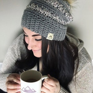 May include: A woman wearing a gray and white crocheted beanie with a faux fur pom pom. The beanie has a brown leather tag with the text "Taylor Made" on it. She is holding a white mug with a mountain graphic and the text "Taylor Made" on it.