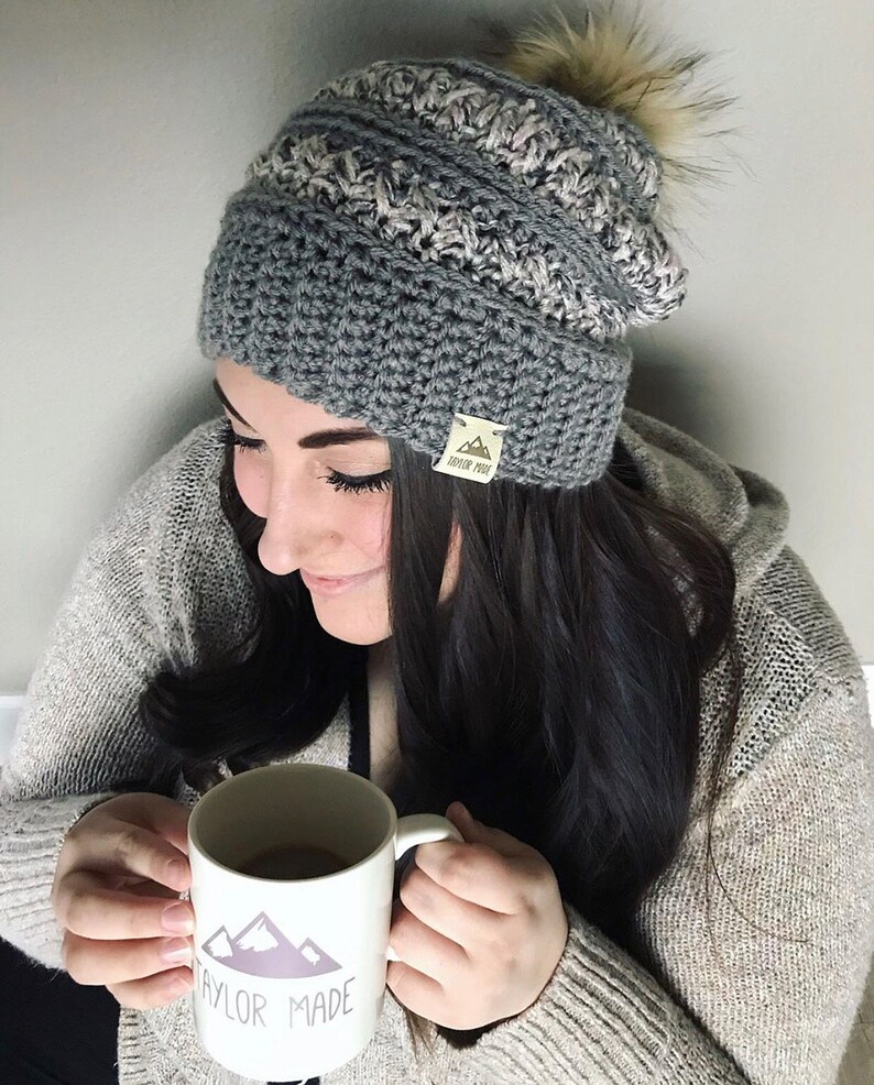 May include: A woman wearing a gray and white crocheted beanie with a faux fur pom pom. The beanie has a brown leather tag with the text "Taylor Made" on it. She is holding a white mug with a mountain graphic and the text "Taylor Made" on it.