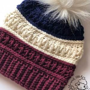 May include: A crocheted beanie hat with a white pom pom. The hat is striped with burgundy, cream, and navy blue yarn. The hat has a label that says "Sheepish Stitches" with a cartoon sheep holding yarn and knitting needles.