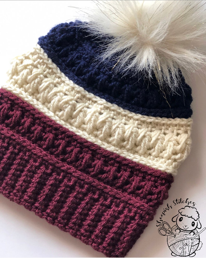 May include: A crocheted beanie hat with a white pom pom. The hat is striped with burgundy, cream, and navy blue yarn. The hat has a label that says "Sheepish Stitches" with a cartoon sheep holding yarn and knitting needles.