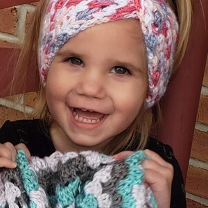 May include: A young girl wearing a pink, white, and gray crocheted headband. She is smiling and holding a teal, white, and gray crocheted blanket.