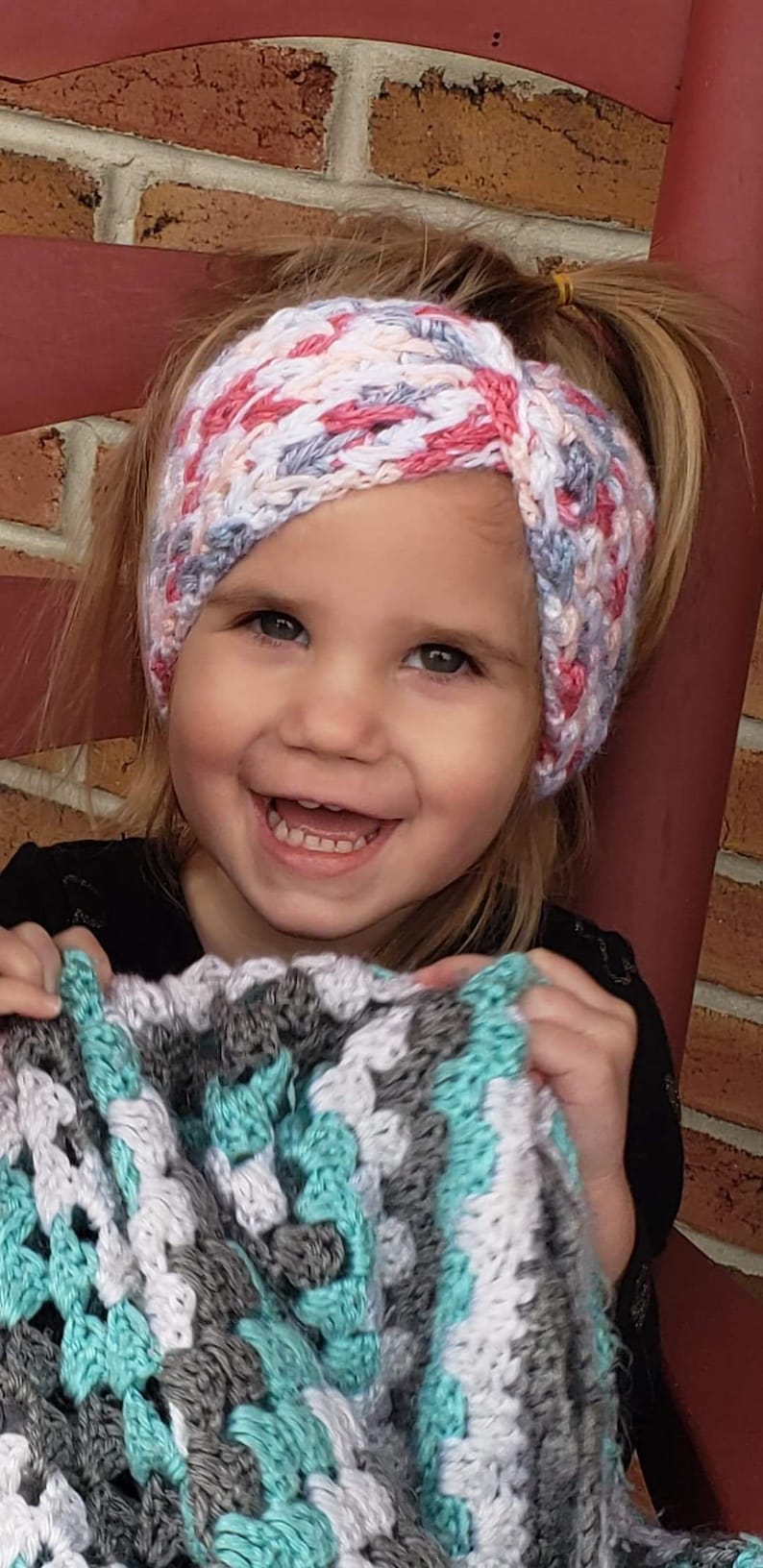 May include: A young girl wearing a pink, white, and gray crocheted headband. She is smiling and holding a teal, white, and gray crocheted blanket.