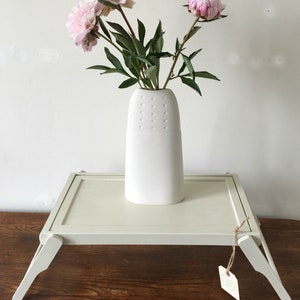 May include: A white folding tray table with a white ceramic vase with pink peonies on top. The tray table has a light wood grain finish and is sitting on a wooden surface.