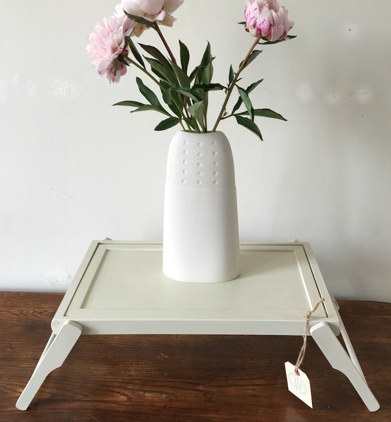 May include: A white folding tray table with a white ceramic vase with pink peonies on top. The tray table has a light wood grain finish and is sitting on a wooden surface.