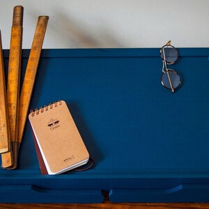 May include: A blue wooden easel with a small brown notebook that says "Grain" on the cover, three wooden rulers, and a pair of gold-rimmed glasses.