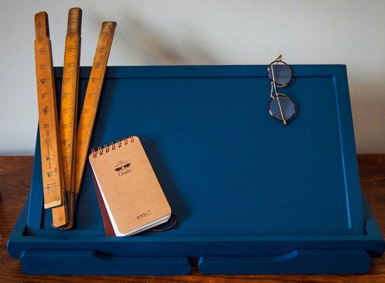 May include: A blue wooden easel with a small brown notebook that says "Grain" on the cover, three wooden rulers, and a pair of gold-rimmed glasses.
