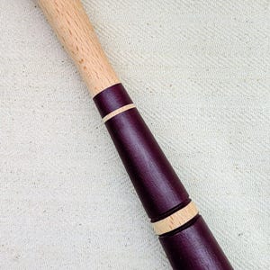 May include: A wooden muddler with a light-coloured handle and a dark purple body. The muddler has a rounded end for crushing ingredients and is accented with light wood rings.