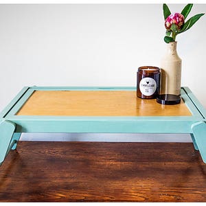 May include: A light blue wooden tray table with a light brown top. The table has two foldable legs and is decorated with a candle and a vase with a pink flower.