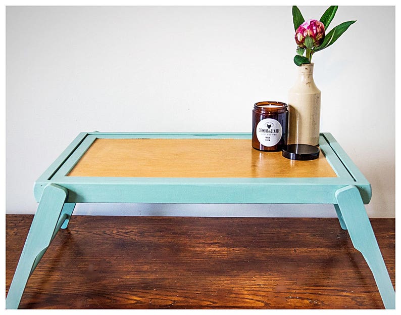 May include: A light blue wooden tray table with a light brown top. The table has two foldable legs and is decorated with a candle and a vase with a pink flower.