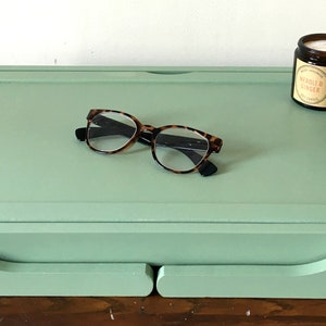 May include: A light green wooden tray with a raised edge and two drawers. The tray has a pair of eyeglasses and a small candle jar on top. The candle jar has a label that reads "Vanilla Ginger".