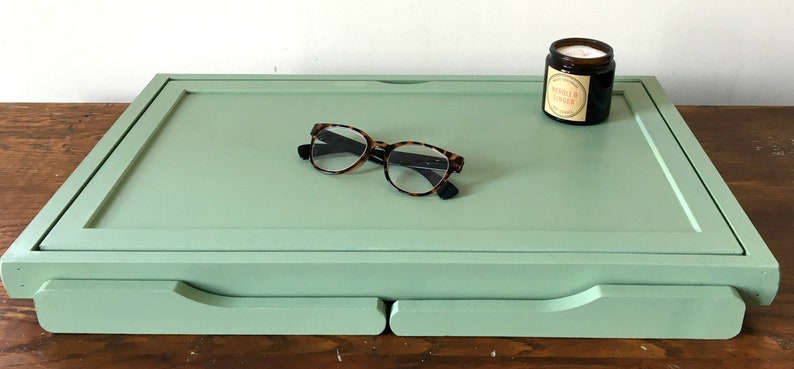 May include: A light green wooden tray with a raised edge and two drawers. The tray has a pair of eyeglasses and a small candle jar on top. The candle jar has a label that reads "Vanilla Ginger".