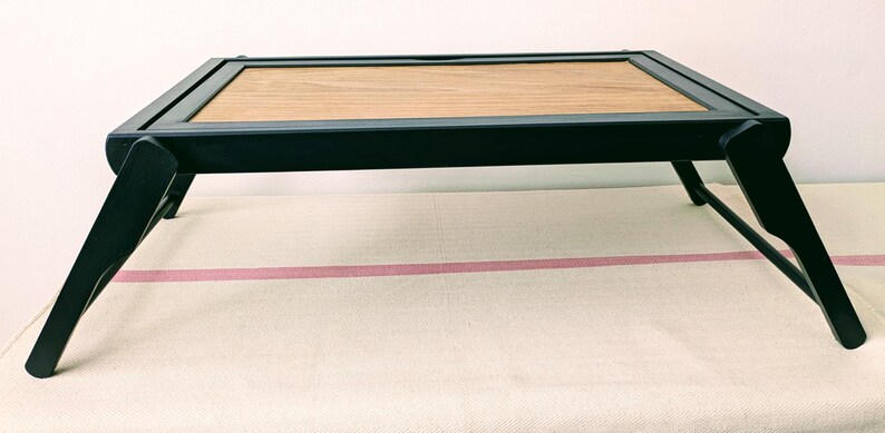 May include: A black wooden folding tray with a rectangular wooden top. The tray is folded and sitting on a white surface.