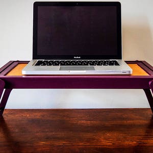 May include: A purple laptop stand with a laptop on top. The stand is made of wood and has a foldable design. The laptop is a silver MacBook with a black screen.