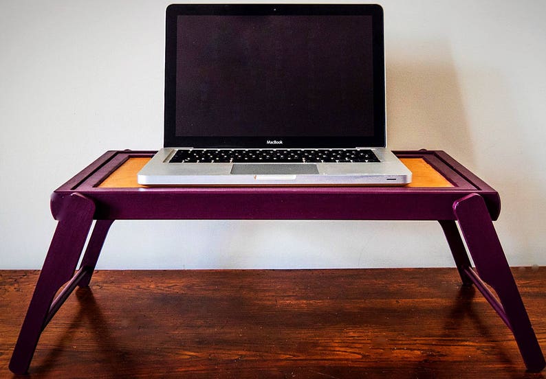 May include: A purple laptop stand with a laptop on top. The stand is made of wood and has a foldable design. The laptop is a silver MacBook with a black screen.