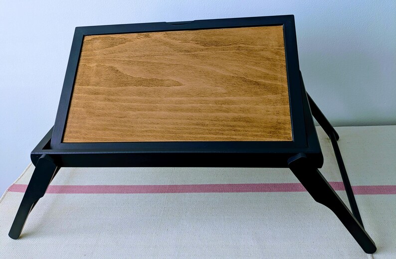 May include: A black wooden folding tray with a brown wood top. The tray is folded and sitting on a white and pink striped surface.