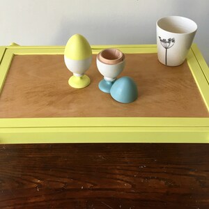 May include: A yellow tray table with a wooden top and yellow legs. The tray table has a white ceramic cup with a black flower design on it. There are three wooden eggs on the tray table, one yellow and white, one white with a wooden top, and one blue.