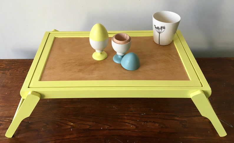 May include: A yellow tray table with a wooden top and yellow legs. The tray table has a white ceramic cup with a black flower design on it. There are three wooden eggs on the tray table, one yellow and white, one white with a wooden top, and one blue.