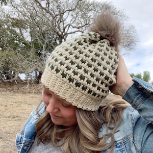 以下が含まれることがあります： ベージュとグリーンのクロシェ編みビーニーにフェイクファーのポンポンが付いている。ビーニーはテクスチャのあるステッチパターンになっている。