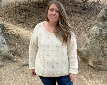 May include: A woman wearing a white knitted sweater with a delicate floral pattern, blue jeans, and brown knee-high boots. She is standing in a natural outdoor setting.