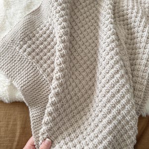 May include: A beige knitted blanket with a textured pattern. The blanket has a thick, ribbed border and a diamond-like pattern across the main body. The blanket is folded, revealing the texture and color variations. The blanket is soft and cozy.