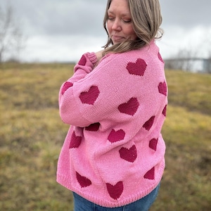 May include: A pink knitted jumper with a heart pattern in a darker shade of pink. The jumper is being worn by a person with long brown hair.
