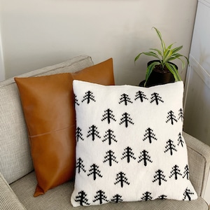 Op de afbeelding: Een wit kussen met een zwart gebreid patroon van dennenbomen. Het kussen ligt op een bruine leren bank met een wit en grijs gestreept patroon.
