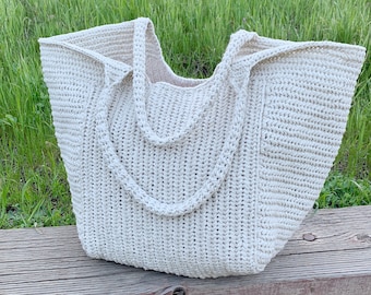 Puede incluir: Un bolso tote grande de crochet de color blanco roto con asas dobles gruesas. El bolso está sobre una superficie de madera con hierba verde en el fondo.