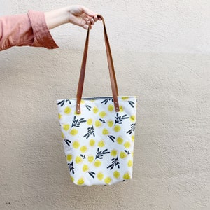 Könnte beinhalten: Eine weiße Einkaufstasche mit einem Blumenmuster aus gelben Blüten und schwarzen Stielen. Die Tasche hat braune Lederriemen und einen Reißverschluss.