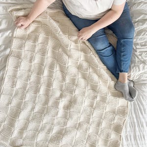 Puede incluir: Una manta de color crema tejida a mano con un patrón tejido texturizado. La manta es rectangular con un borde festoneado. La manta está sobre una cama blanca, y una persona es parcialmente visible, sosteniendo la manta.