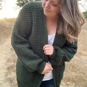 Pode incluir: Um cardigan de malha verde escuro com um caimento relaxado e mangas compridas. O cardigan é aberto na frente e tem um padrão de malha texturizado.