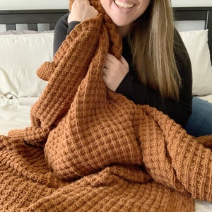 May include: A brown crocheted blanket with a waffle stitch pattern. The blanket is folded and draped over a bed.