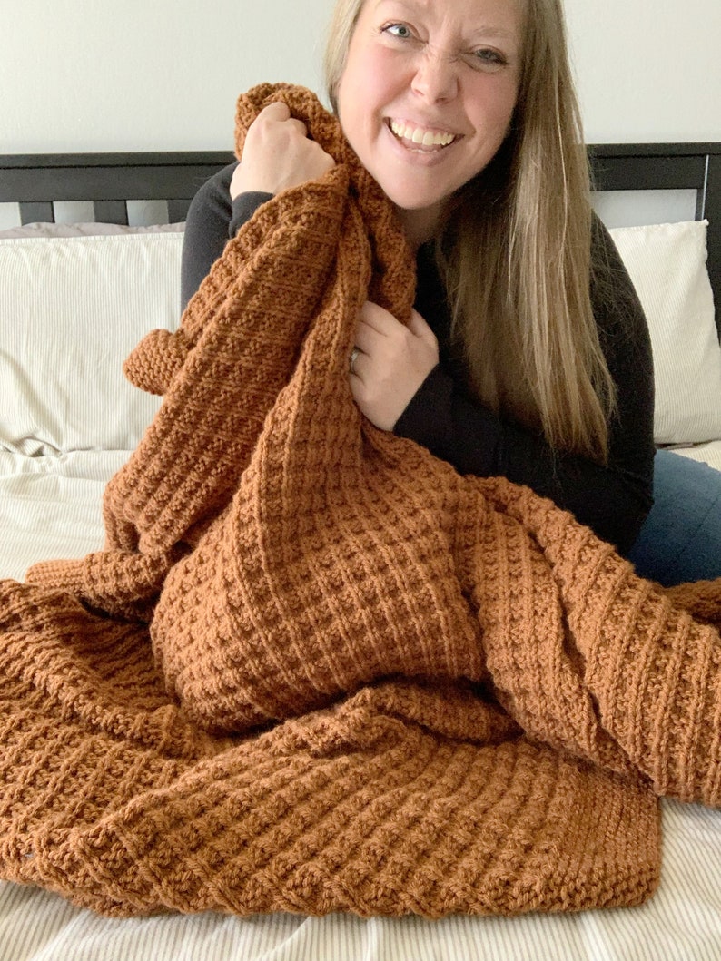 May include: A brown crocheted blanket with a waffle stitch pattern. The blanket is folded and draped over a bed.