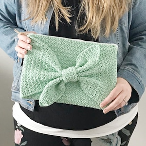 May include: A light green crocheted clutch with a large bow in the center. The clutch is held by a person wearing a denim jacket.