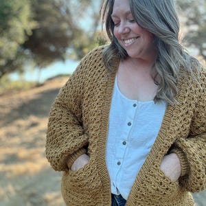 以下が含まれることがあります： マスタードイエローのカーディガン。質感のあるニットの模様とポケットが付いています。カーディガンはフロントが開いており、ゆったりとしたフィット感です。