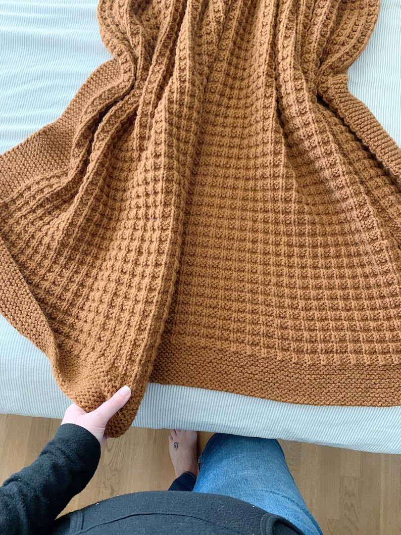 May include: A brown knitted throw blanket with a waffle stitch pattern. The blanket is folded in half and is being held by a hand.
