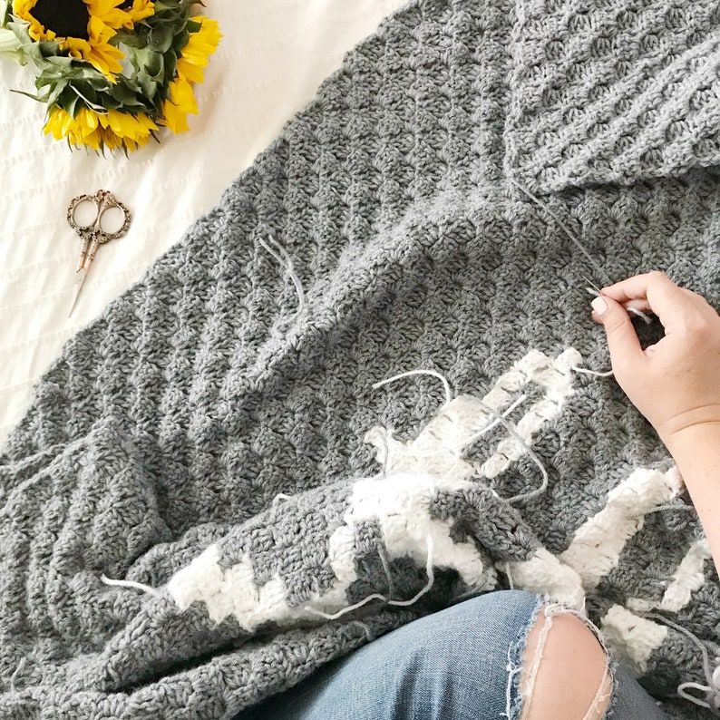 K&ouml;nnte beinhalten: Eine graue und wei&szlig;e H&auml;keldecke mit einem strukturierten Muster. Die Decke wird mit wei&szlig;em Garn und einer H&auml;kelnadel bearbeitet.