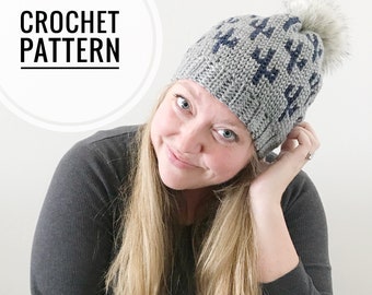 May include: A gray and navy blue crochet beanie with a white pom pom. The beanie has a repeating pattern of small triangles. The beanie is being worn by a person with long blonde hair.
