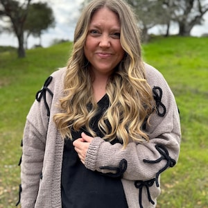 May include: A light brown cardigan with black bow accents on the sleeves and sides. The open-front sweater is worn over a black top. The woman in the image is smiling, with long blonde hair, and is standing outside in front of a green background.