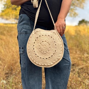 May include: A round, beige crochet shoulder bag featuring a textured, circular pattern with a central medallion and a long, matching crocheted strap. The bag is crafted from a light-colored yarn and displays a consistent, intricate stitch design.
