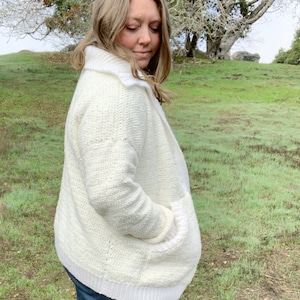 May include: A white crocheted cardigan with a white collar and pockets. The cardigan is worn by a person standing in a grassy field.