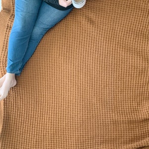 May include: A person sitting on a brown waffle knit throw blanket. The blanket has a textured waffle weave pattern. The person is holding a white mug.