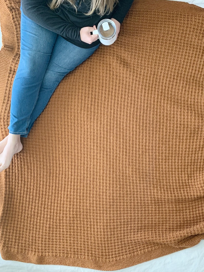 May include: A person sitting on a brown waffle knit throw blanket. The blanket has a textured waffle weave pattern. The person is holding a white mug.