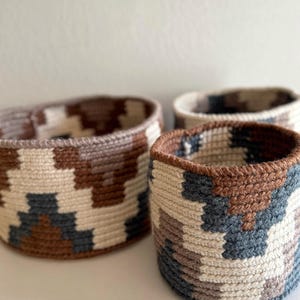 May include: Three handwoven baskets with geometric patterns in shades of brown, cream, and gray. The baskets are cylindrical with a slightly flared top. The texture of the woven material is visible, adding to the handmade aesthetic. These baskets are suitable for storage or decoration.
