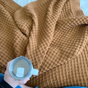 May include: A person sitting on a bed with a brown knitted blanket and holding a white travel mug with a lid.