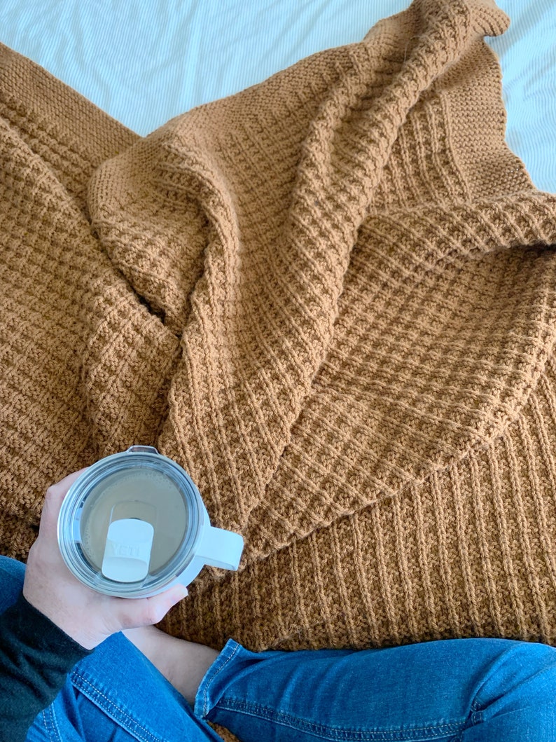 May include: A person sitting on a bed with a brown knitted blanket and holding a white travel mug with a lid.