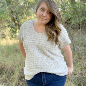 May include: A woman wearing a white crocheted top with a textured pattern and blue jeans.