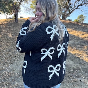 May include: A black sweater with white bow patterns. The sweater is a dark color and features multiple white bows across the front and back. The sweater is made of a thick knit material.