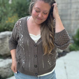 May include: A brown crochet cardigan with a floral design and button closure. The cardigan is worn over a white top and paired with blue jeans. The sleeves are three-quarter length.