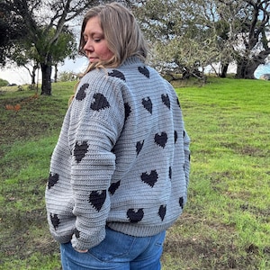 May include: A gray, knitted cardigan with black heart-shaped patterns. The sweater has long sleeves and a ribbed texture. The person is wearing blue jeans. The background features green grass and trees.